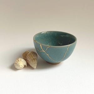 May include: A teal ceramic bowl with gold kintsugi repair lines. Two seashells sit beside the bowl. The bowl has a smooth, glossy finish and an irregular shape. The kintsugi technique highlights the bowl's history and adds a decorative element.