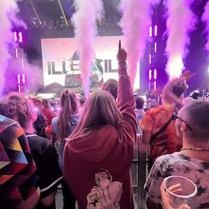 May include: A crowd of people at a concert with a large screen displaying the word "ILLENIUM" in white letters on a blue and green background. The crowd is cheering and waving their hands in the air. There is a lot of smoke and fog in the air.
