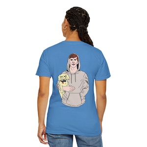 May include: A blue t-shirt with a graphic of a person in a grey hoodie holding a small dog. The person is looking to the left and the dog is looking to the right.