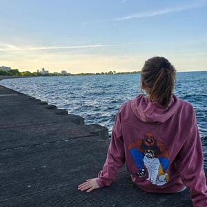 May include: A person wearing a maroon hoodie with a graphic print of a cartoon man holding a dog. The person is sitting on a concrete pier overlooking a body of water.