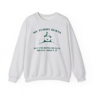 Puede incluir: Una sudadera gris claro con el texto "My tummy hurts but I'm being really brave about it" en verde. Un oso de dibujos animados con gorro de dormir está en una cama con una manta.