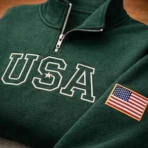 Peut inclure: Un sweat-shirt vert foncé avec un col zippé. Le mot "USA" est brodé en lettres blanches sur la poitrine, avec une étoile dans le "A". Un écusson du drapeau américain est sur la manche.