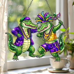Op de afbeelding: Twee groene alligatorfiguren in Mardi Gras-outfit, hangend aan een ketting. Eén draagt een paarse jas en hoed, de ander een gevederde hoofdtooi en houdt een masker vast. Beide hebben kralenkettingen en staan in een danshouding.