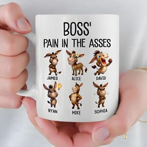 May include: White ceramic mug featuring the text "BOSS' PAIN IN THE ASSES" above cartoon donkeys with names. The donkeys have various expressions. The mug is held by a person.