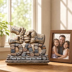 May include: A stone-like sculpture with the word "DAD" in mosaic-style letters. Above, two fists bump, with the names "Olivia" and "Liam" on one, and "Dad" on the other. The base reads "Thank You For Being Our Rock."