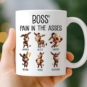 May include: White ceramic mug with the text "BOSS' PAIN IN THE ASSES" in black. The mug features cartoon donkeys with names like James, Alice, and David. The mug is held by a person.