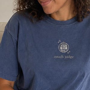 May include: A blue short-sleeve t-shirt featuring a small graphic of a disco ball with sparkles above the text "couch judge" printed in white on the centre chest. The shirt has a crew neckline and a relaxed fit, paired with light blue denim jeans.