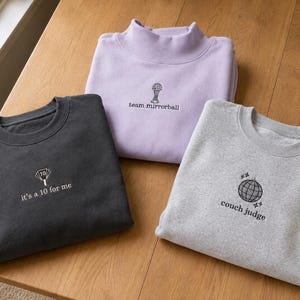 May include: Three crewneck sweatshirts in charcoal, light purple, and heather gray. The charcoal sweatshirt features an embroidered fan with the text 'it's a 10 for me'. The purple one shows a mirrorball with 'team mirrorball', and the gray one has a disco ball with 'couch judge'.