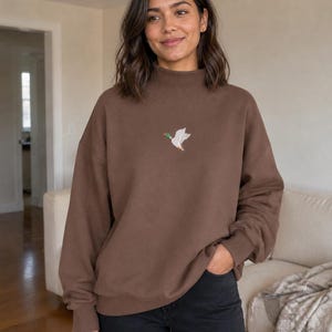 Puede incluir: Una sudadera marrón de manga larga con cuello alto y un pequeño gráfico bordado de un pato real volando en el centro del pecho. La sudadera cuenta con puños y dobladillo acanalados, combinada con unos pantalones vaqueros de color negro.