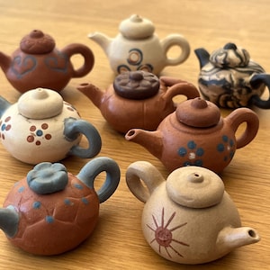 May include: A collection of small, decorative teapots in various colors and designs. The teapots are made of ceramic and feature intricate details, including floral and geometric patterns. The teapots are approximately 3 inches tall.