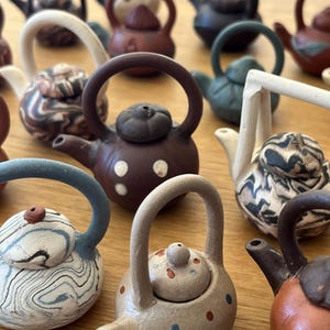 May include: A collection of miniature ceramic teapots in various colors and patterns. The teapots have rounded bodies, small spouts, and arched handles. Some feature speckled designs, while others have swirl or floral patterns. The teapots are arranged on a wooden surface.