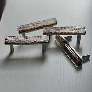 May include: Four rectangular cabinet pulls with a textured, clear glass center and a gold-toned accent. The pulls have a brushed metal base and are arranged on a light gray surface. These decorative pulls are ideal for updating furniture.