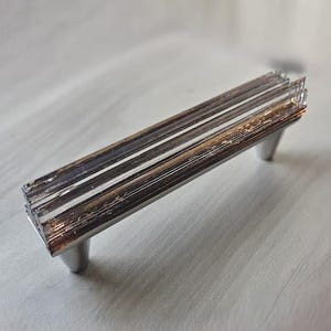 May include: A silver-toned cabinet pull with a rectangular design. The pull features a series of parallel bars with a distressed finish, mounted on two small, rounded feet. The hardware is designed for furniture and cabinetry.