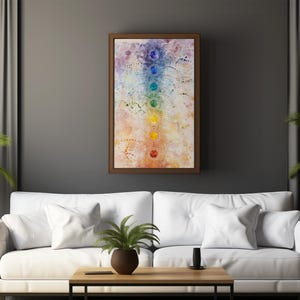 May include: A white couch with white pillows in a living room setting. A framed painting of a colorful chakra symbol hangs on the wall above the couch. A small potted plant sits on the coffee table in front of the couch.