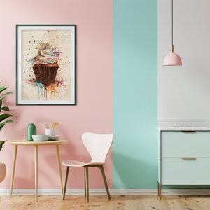 May include: A framed print of a watercolour painting of a cupcake with colourful frosting and sprinkles. The cupcake is on a white background with splatters of paint in various colours. The frame is dark green and the print is hanging on a pink wall.