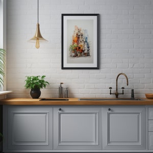 May include: A framed watercolor print of a colorful cityscape, featuring buildings in shades of orange, red, and blue. The artwork is displayed on a white brick wall above a kitchen counter with a plant and a gold-toned light fixture.