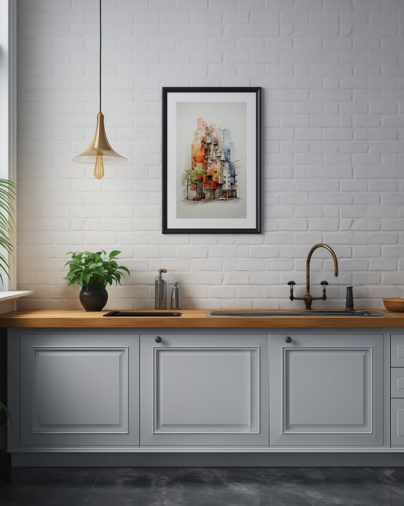 May include: A framed watercolor print of a colorful cityscape, featuring buildings in shades of orange, red, and blue. The artwork is displayed on a white brick wall above a kitchen counter with a plant and a gold-toned light fixture.