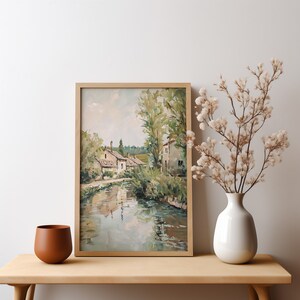 May include: A framed painting of a rural landscape with a river, houses, and trees. The painting is in a light wood frame and has a muted color palette.