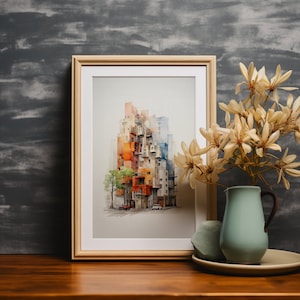 May include: A framed watercolor print of a colorful cityscape, featuring buildings in shades of orange, red, and blue. The artwork is displayed on a wooden table, next to a light blue pitcher with dried flowers and a decorative stone.