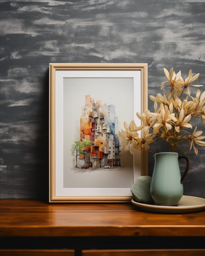 May include: A framed watercolor print of a colorful cityscape, featuring buildings in shades of orange, red, and blue. The artwork is displayed on a wooden table, next to a light blue pitcher with dried flowers and a decorative stone.
