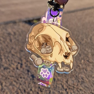 Könnte beinhalten: Ein durchsichtiger Acryl-Schlüsselanhänger mit einem Cartoon-Katzenkopf und einem Pfoten-Anhänger. Der Schädel ist mit kleinen Metallschrauben und Muttern gefüllt.