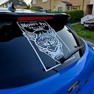 Puede incluir: Un coche azul con una pegatina blanca en la ventana trasera. La pegatina presenta una ilustración en blanco y negro de un demonio con cuernos y el texto "MONEY PIT" y "マニー・ピット" en japonés. El texto "Double Eclipse" también está en la pegatina.