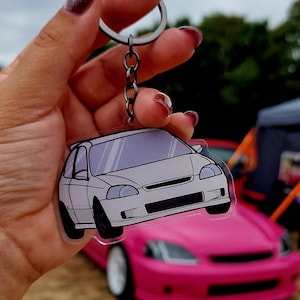 Pode incluir: Um porta-chaves de acrílico transparente com um carro hatchback ilustrado em branco com rodas pretas e uma perspetiva frontal. O porta-chaves está preso a um anel de metal prateado e a uma corrente curta, exibindo um design gráfico detalhado.