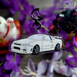 Op de afbeelding: Een witte autosleutelhanger met zwarte wielen en een zwarte sluiting. De sleutelhanger hangt voor paarse bloemen en een vage achtergrond met een gelukskatfiguur.