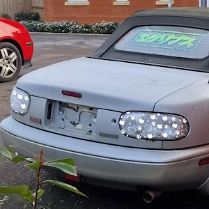 May include: Silver convertible car with custom cat paw and cat face taillight designs. The rear window has a graphic that says "Eclipse". A red car is partially visible in the background.
