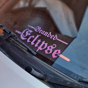 Puede incluir: Una calcomanía de vinilo rosa y púrpura con el texto "Branded Eclipse" en el parabrisas de un coche blanco.