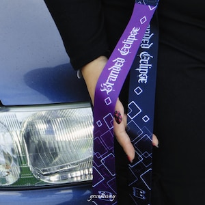 Może przedstawiać: Dwie smycze z napisem "Branded Eclipse" w białym skrypcie. Jedna smycz jest fioletowa z białymi geometrycznymi kształtami, a druga granatowa z białymi geometrycznymi kształtami. Smycze są trzymane przed niebieskim samochodem.