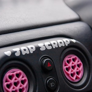 May include: A black car interior with a white sticker that says "Jap Scrap" and two pink heart-shaped air freshener vents.