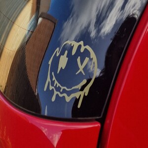 Puede incluir: Un coche rojo con una pegatina de cara sonriente dorada en la ventana trasera. La pegatina tiene un efecto de goteo y los ojos están tachados.