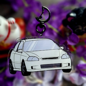 May include: White car-shaped keyring with black wheels and a black metal clasp. The car has a detailed design with windows and a front grill. The background is blurred with purple flowers and other objects.