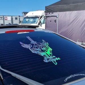 Könnte beinhalten: Ein silbernes Auto mit einem Aufkleber an der Heckscheibe, der ein grünes und blaues holografisches Design eines Dämonenkopfes mit einem Schwert zeigt. Der Aufkleber lautet "Branded Eclipse" und "Caffeine & Machine".