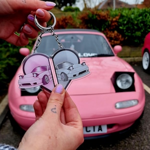 May include: A pink car with a heart-shaped keyring featuring two cartoon cars. The keyring is made of clear acrylic and has a silver metal ring.