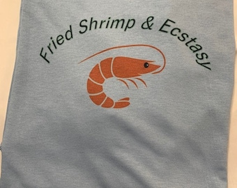 May include: Light blue t-shirt with the words "Fried Shrimp & Ecstasy" in a curved font above an orange shrimp graphic. The shirt is folded and appears to be made of a soft material.