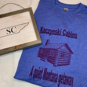 May include: A blue t-shirt with the text "Kaczynski Cabins" and "A quiet Montana getaway" in purple. A purple illustration of a log cabin is centered on the shirt.