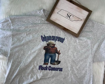 May include: A heather gray t-shirt featuring a graphic of a brown bear wearing a hat and blue jeans while holding a shovel. The text above the bear reads "Only you can prevent" and below it reads "Flock Cameras". A wooden sign with "SC" is present.