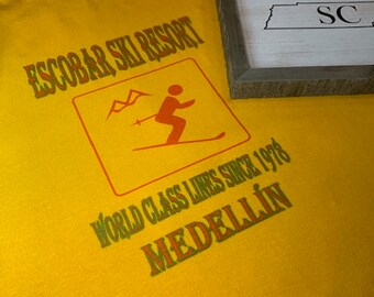 May include: Yellow t-shirt with a green and orange graphic that says "Escobar Ski Resort" and "World Class Lines Since 1978 Medellin".