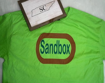 May include: A lime green t-shirt with a brown and teal oval design that says "Sandbox". A small wooden framed sign with the letters "SC" and a state outline is on top of the shirt.