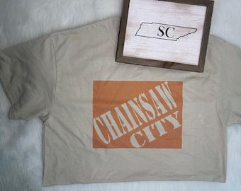 May include: Beige t-shirt with an orange rectangle featuring the words "CHAINSAW CITY" in white. A small wooden framed sign with the outline of Tennessee and the letters "SC" rests on the shirt.