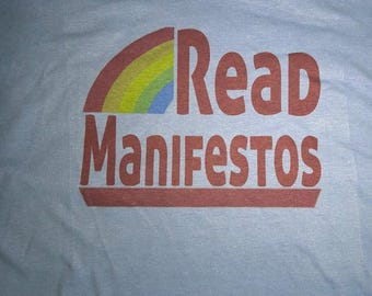 May include: A light blue t-shirt featuring a graphic print with a semi-circular rainbow in red, orange, yellow, green, and blue. Below the rainbow, the text Read Manifestos is printed in a bold, dark red, rounded sans-serif font.