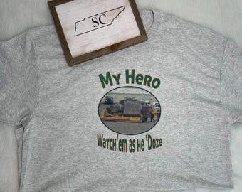 May include: A light gray t-shirt featuring a graphic print with the text "My Hero" and "Watch'em as he 'Doze" in green letters. The center graphic shows a yellow bulldozer. A wooden sign with the letters "SC" inside a state outline is visible.