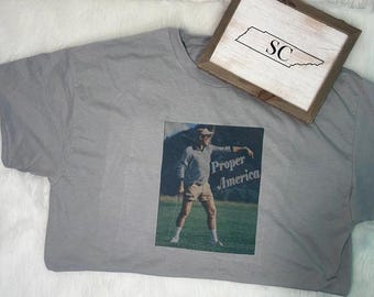 May include: A grey t-shirt featuring a graphic print of a person wearing a cap, long-sleeved shirt, and shorts while standing on a grassy field. The text "Proper America" is printed on the graphic. A wooden framed sign with the letters "SC" is included.