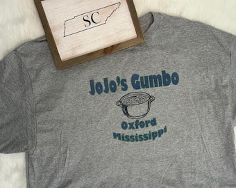 May include: A gray t-shirt with the text "JoJo's Gumbo" above a pot illustration and "Oxford Mississippi" below. A framed wooden sign with the outline of Tennessee and the letters "SC" is in the background.