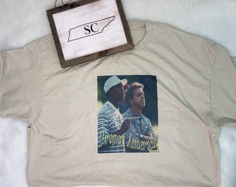 May include: A beige t-shirt featuring a graphic print of two individuals on a golf course, with the text "Proper America" in yellow script below them. A wooden sign with the letters "SC" inside a state outline is positioned above the shirt.