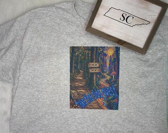 May include: A heather gray t-shirt featuring a colorful graphic of a forest path with a signpost reading This Way and That Way, accompanied by the text Take the Side Quest in blue lettering, positioned below a framed sign with the letters SC.