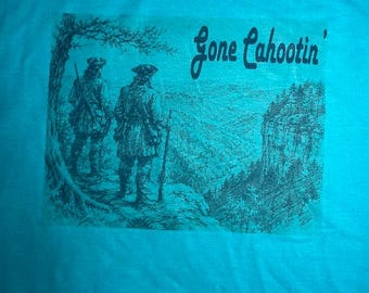 May include: A bright turquoise t-shirt featuring a dark ink-style graphic of two figures in colonial-era clothing standing on a cliff edge overlooking a valley. The text Gone Cahootin' is printed in a cursive script above the landscape scene.
