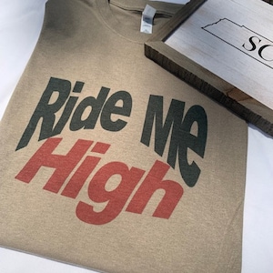 Könnte beinhalten: Ein beiges T-Shirt mit dem Aufdruck "Ride Me High" in schwarzen und roten Buchstaben.